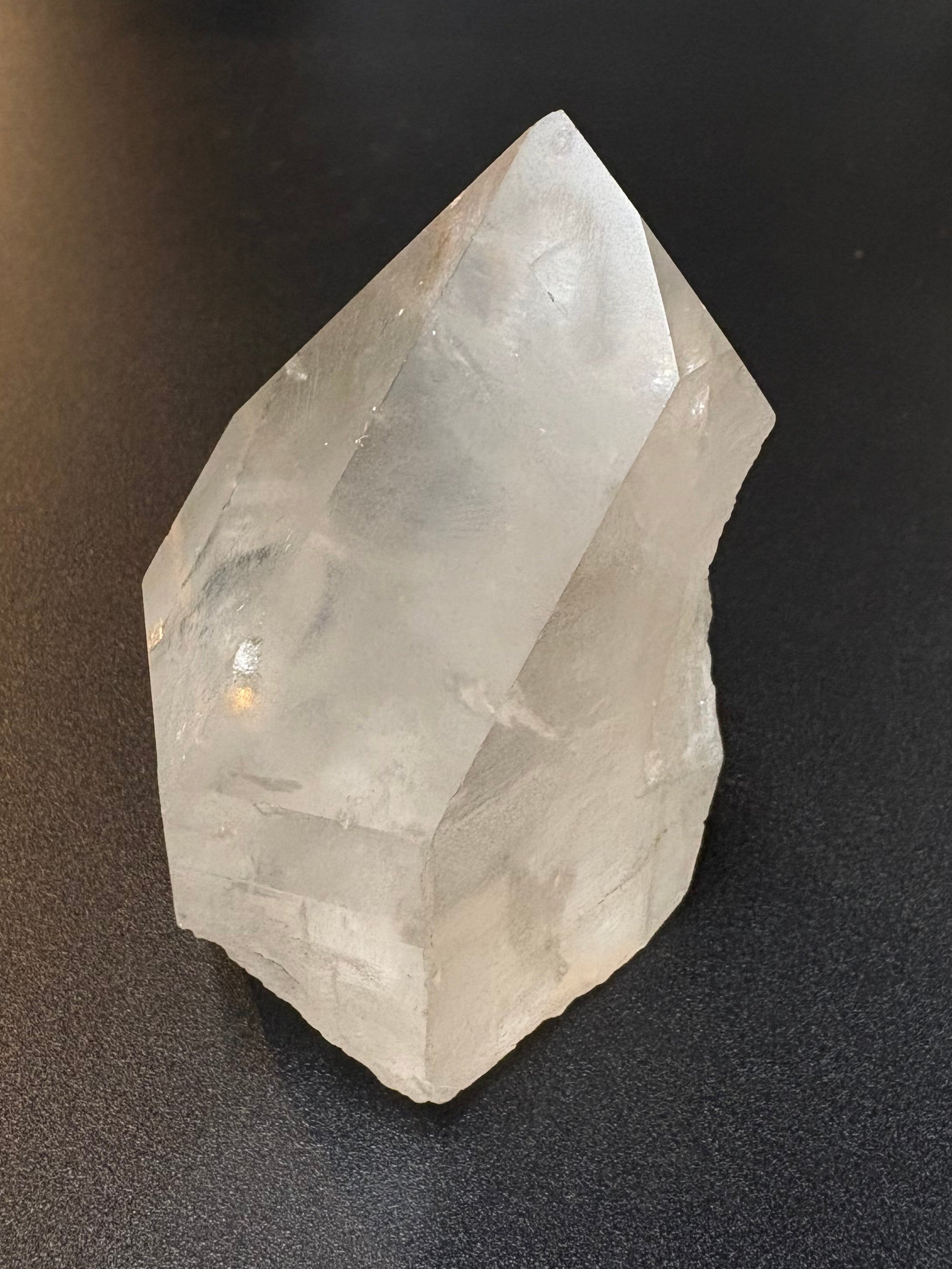 A raw, unpolished clear quartz crystal point resting on a dark gray textured surface.