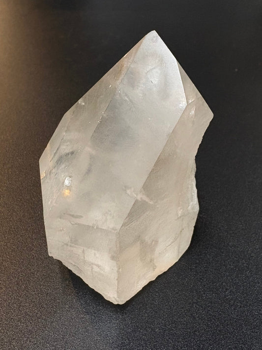 A raw, unpolished clear quartz crystal point resting on a dark gray textured surface.