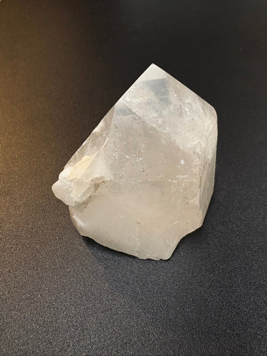 A rough, unpolished translucent white crystal stone with natural jagged edges, resting on a dark textured surface.