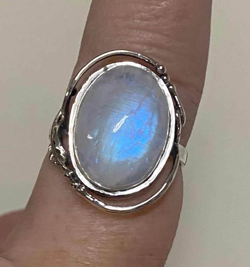 A sterling silver ring with an oval moonstone cabochon, shown worn on a finger. The moonstone displays a blue iridescent sheen, and the silver band has a rustic, hand-hammered texture.