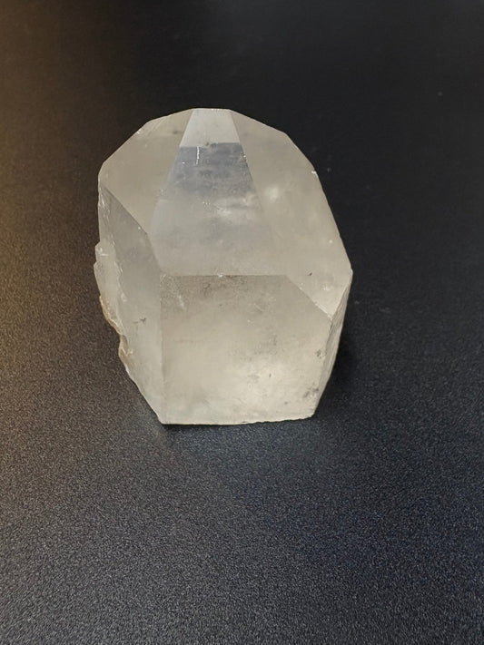 A raw, faceted clear quartz crystal resting on a dark gray textured surface.