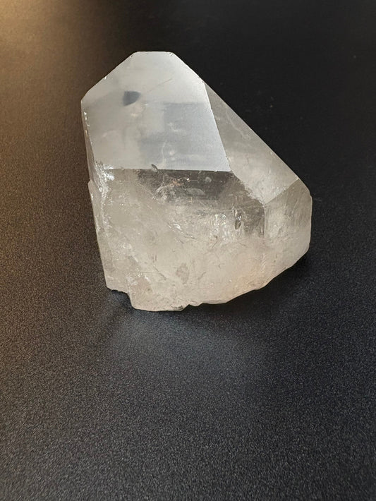 A raw, unpolished clear quartz crystal point resting on a dark gray textured surface.