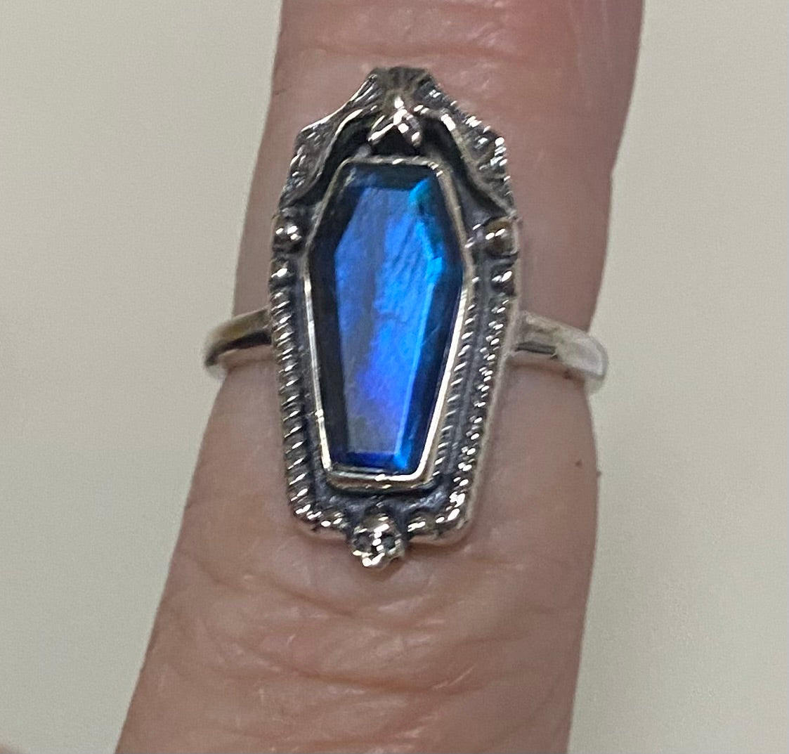 A silver ring with a coffin-shaped blue iridescent gemstone, shown worn on a finger against a plain light background.