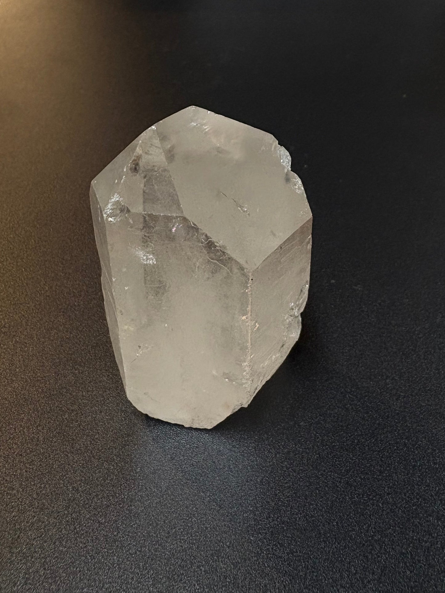 A raw, unpolished clear quartz crystal with natural geometric facets, resting on a dark gray textured surface.