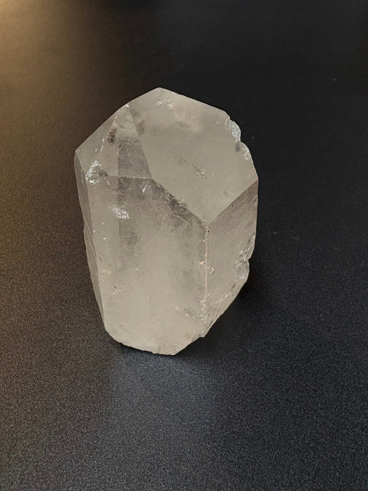 A raw, unpolished clear quartz crystal with natural geometric facets, resting on a dark gray textured surface.
