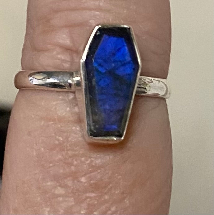 A silver coffin-shaped ring with a vibrant blue labradorite stone, shown worn on a finger.