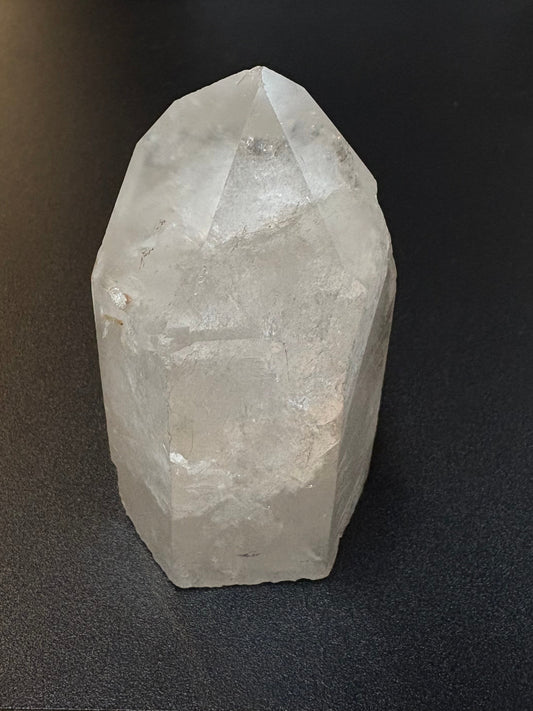 A faceted clear quartz crystal cluster resting on a black textured surface next to a wooden table edge.