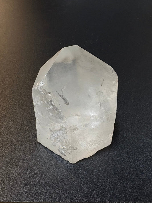 A rough, uncut clear quartz crystal stone resting on a black textured surface next to a wooden table edge.
