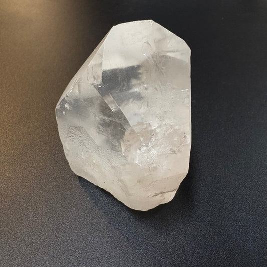 A raw, unpolished clear quartz crystal point with natural facets, resting on a dark textured surface.