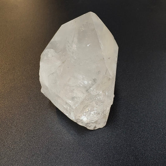 A raw, unpolished clear quartz crystal with natural geometric facets, resting on a dark textured surface.