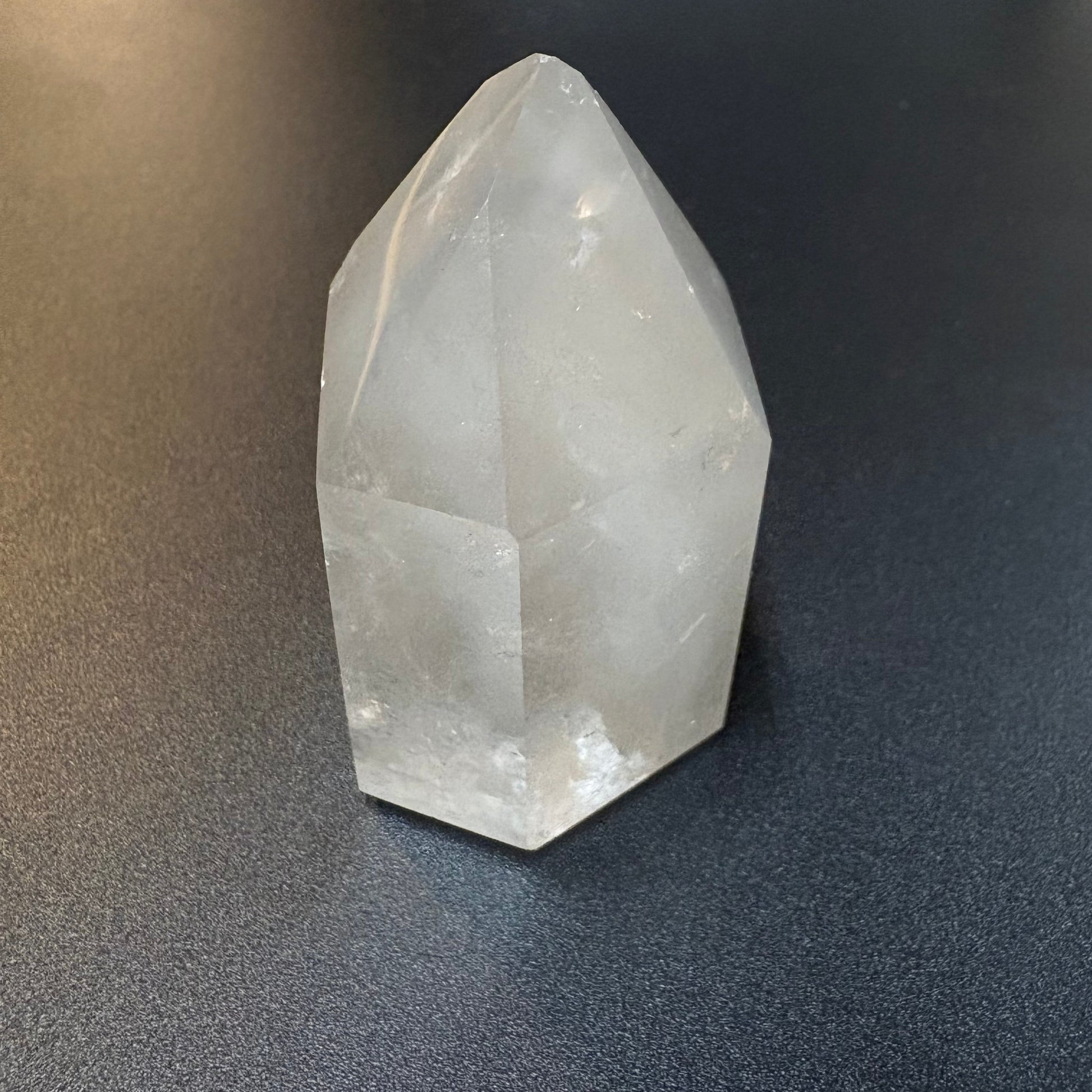 A natural clear quartz crystal point with geometric facets, resting on a dark textured surface.