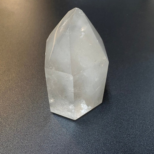 A natural clear quartz crystal point with geometric facets, resting on a dark textured surface.