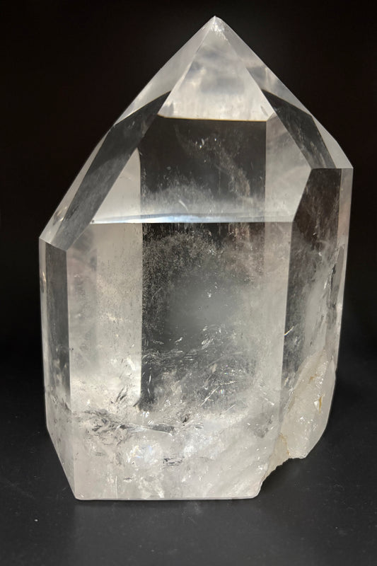 A natural clear quartz crystal point with sharp geometric facets and internal inclusions, photographed against a dark black background.