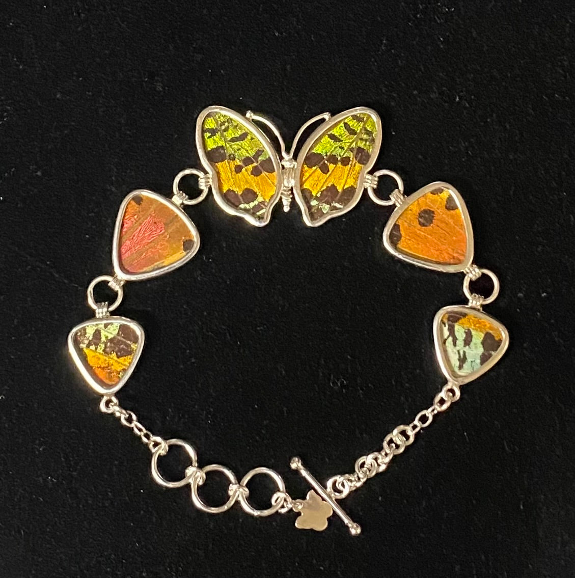 A silver chain bracelet featuring butterfly wings and guitar-pick-shaped inlays with vibrant orange, yellow, and green beetle wing patterns, displayed on a black background.