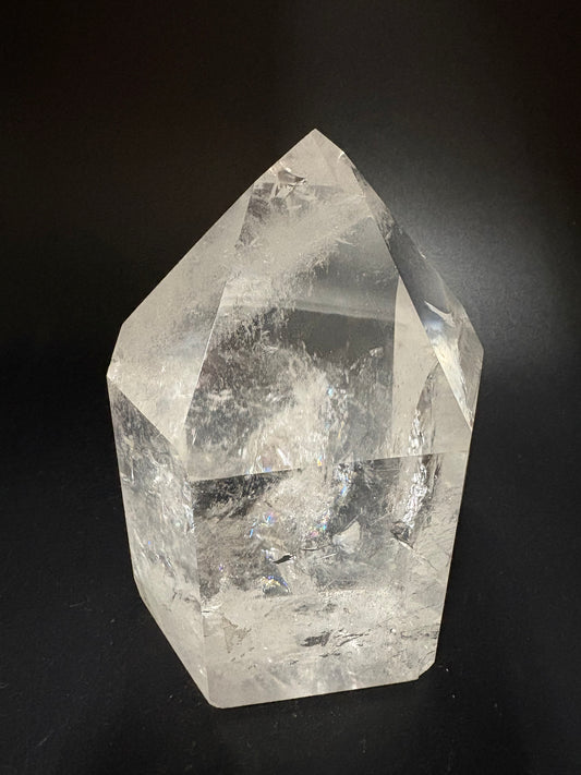 A raw, faceted clear quartz crystal with natural inclusions, displayed on a dark black background.
