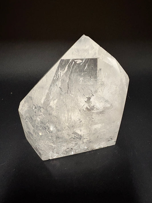 A raw, uncut clear quartz crystal with visible internal inclusions and natural facets, resting on a dark black background.