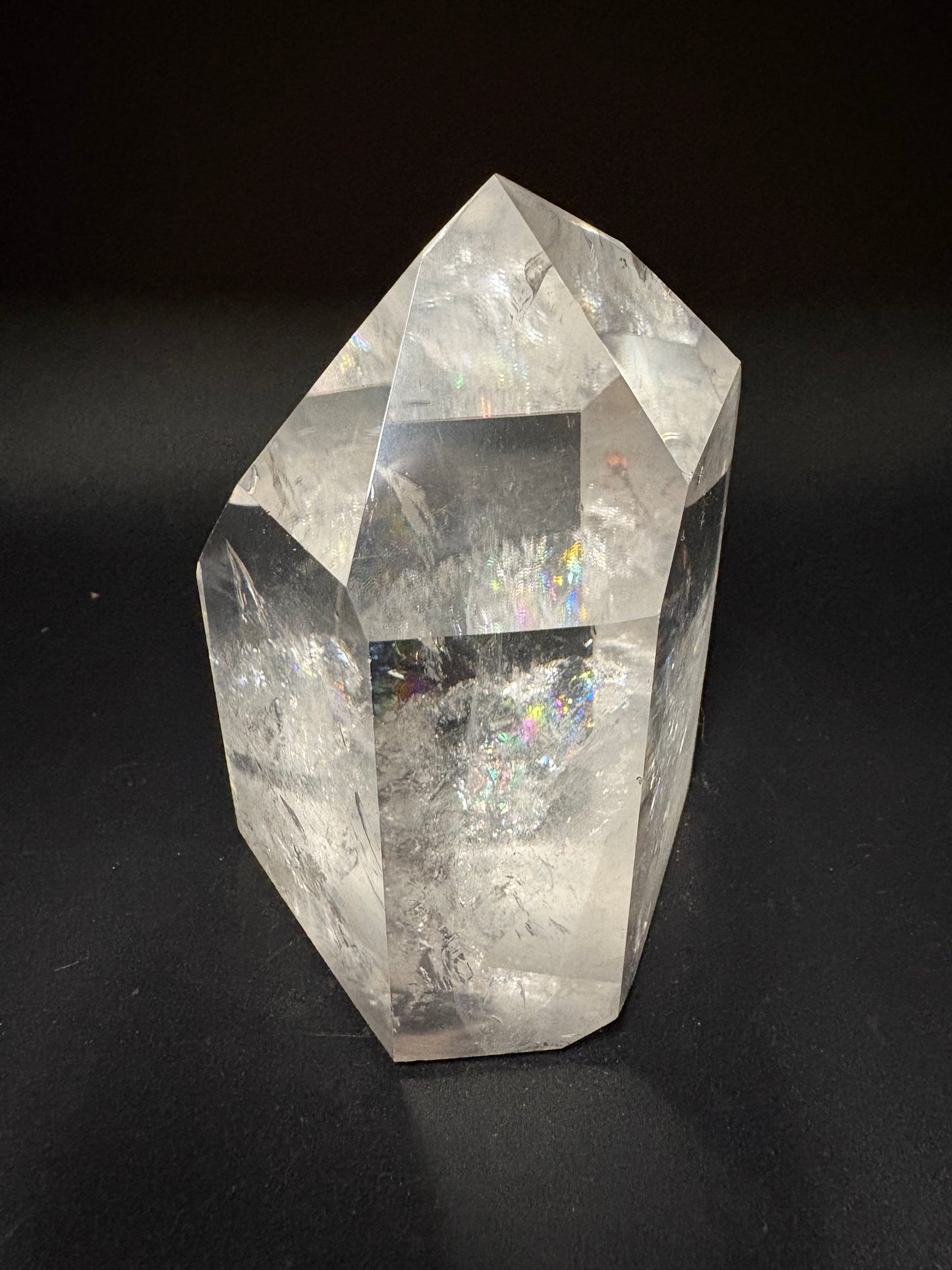 A clear faceted crystal stone with natural internal inclusions and a subtle iridescent sheen, photographed on a dark black background.