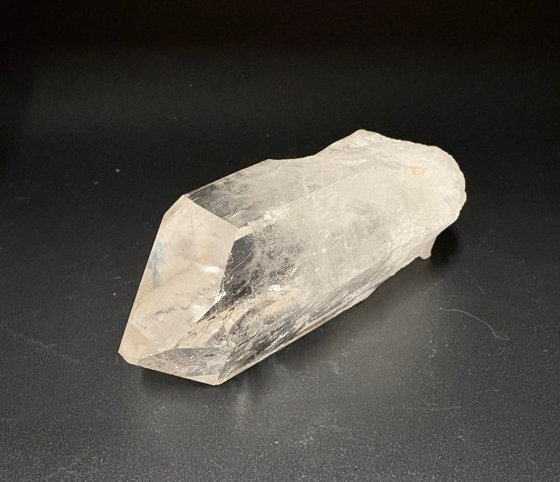 A raw, unpolished clear quartz crystal point resting on a dark, matte surface.