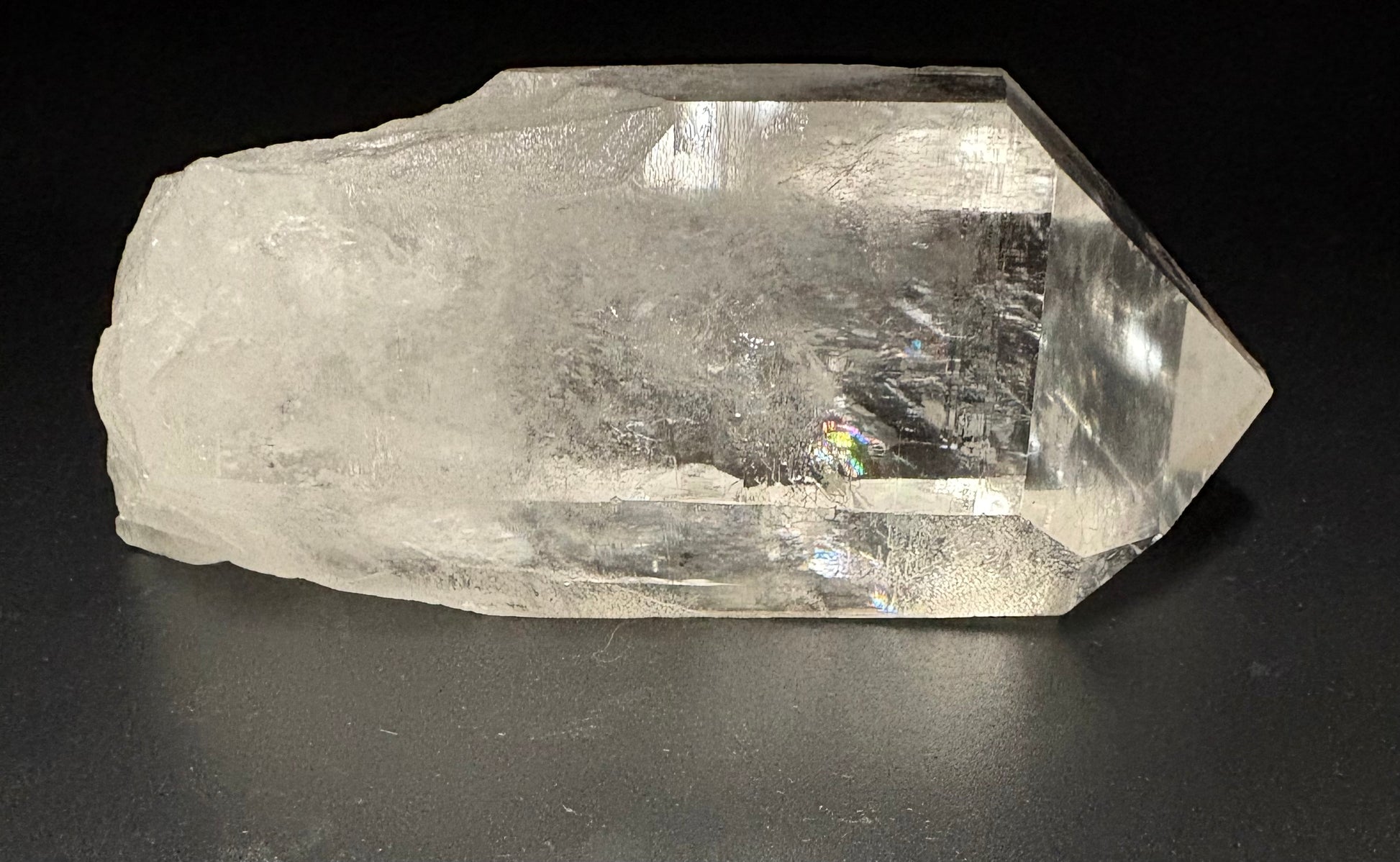 A raw, rough-cut clear quartz crystal point with natural inclusions and a textured, unpolished surface, photographed on a dark gray background.