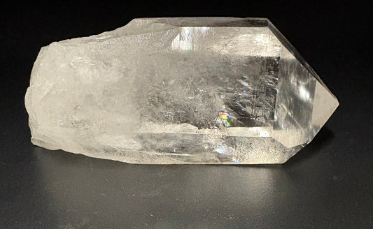 A raw, rough-cut clear quartz crystal point with natural inclusions and a textured, unpolished surface, photographed on a dark gray background.