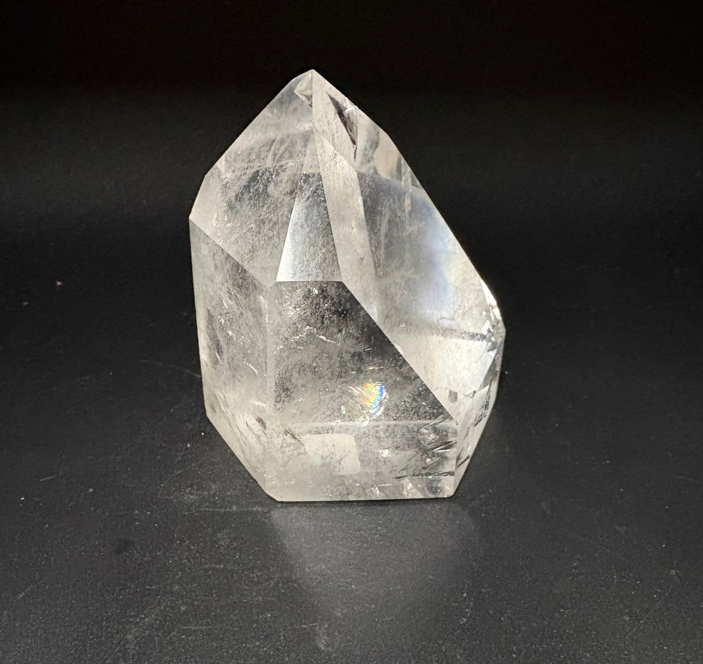A clear quartz crystal point with natural inclusions, standing on a dark black surface against a black background.