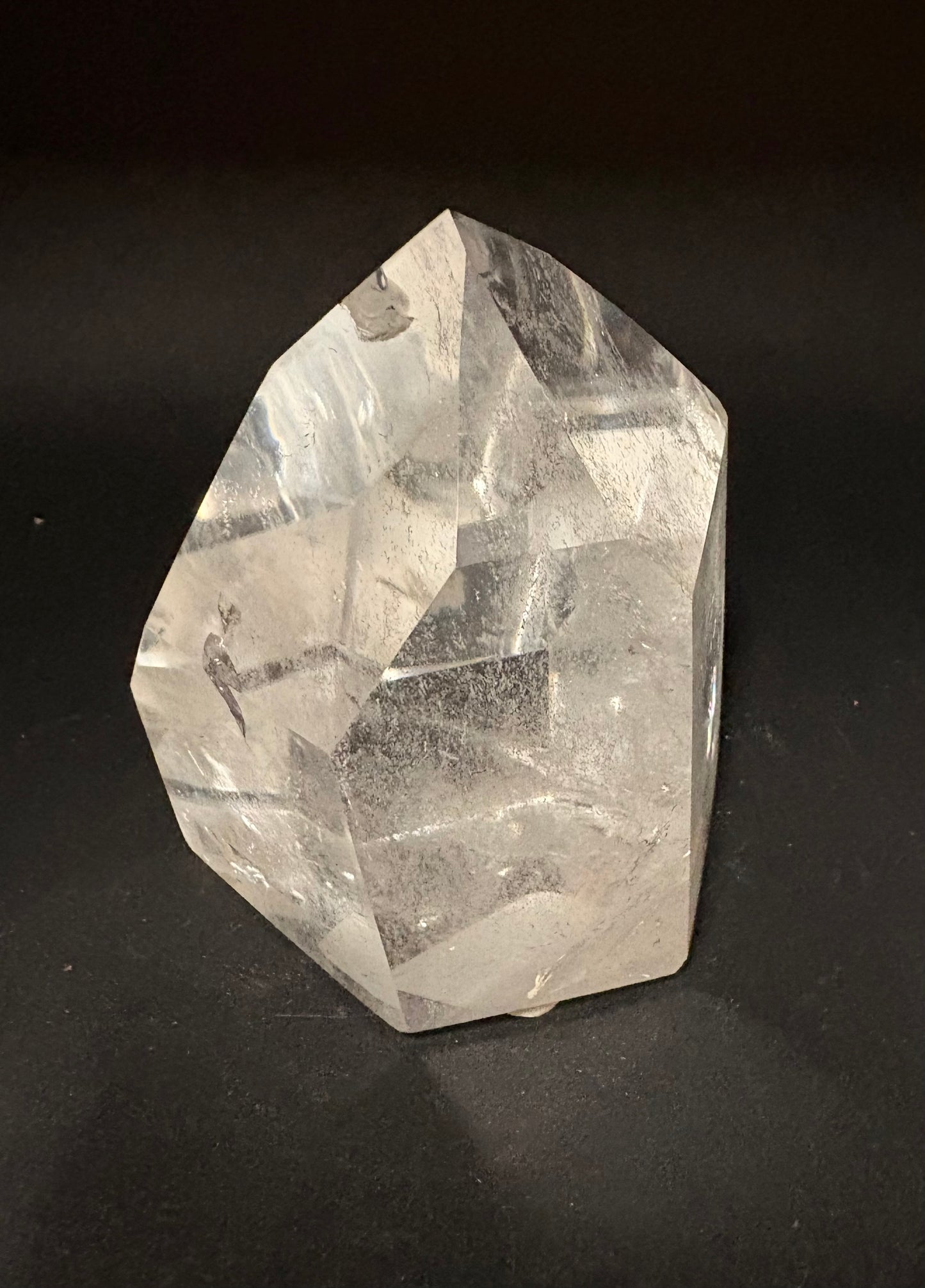 A raw, unpolished clear quartz crystal point with natural faceted edges and internal inclusions, standing upright on a dark black background.