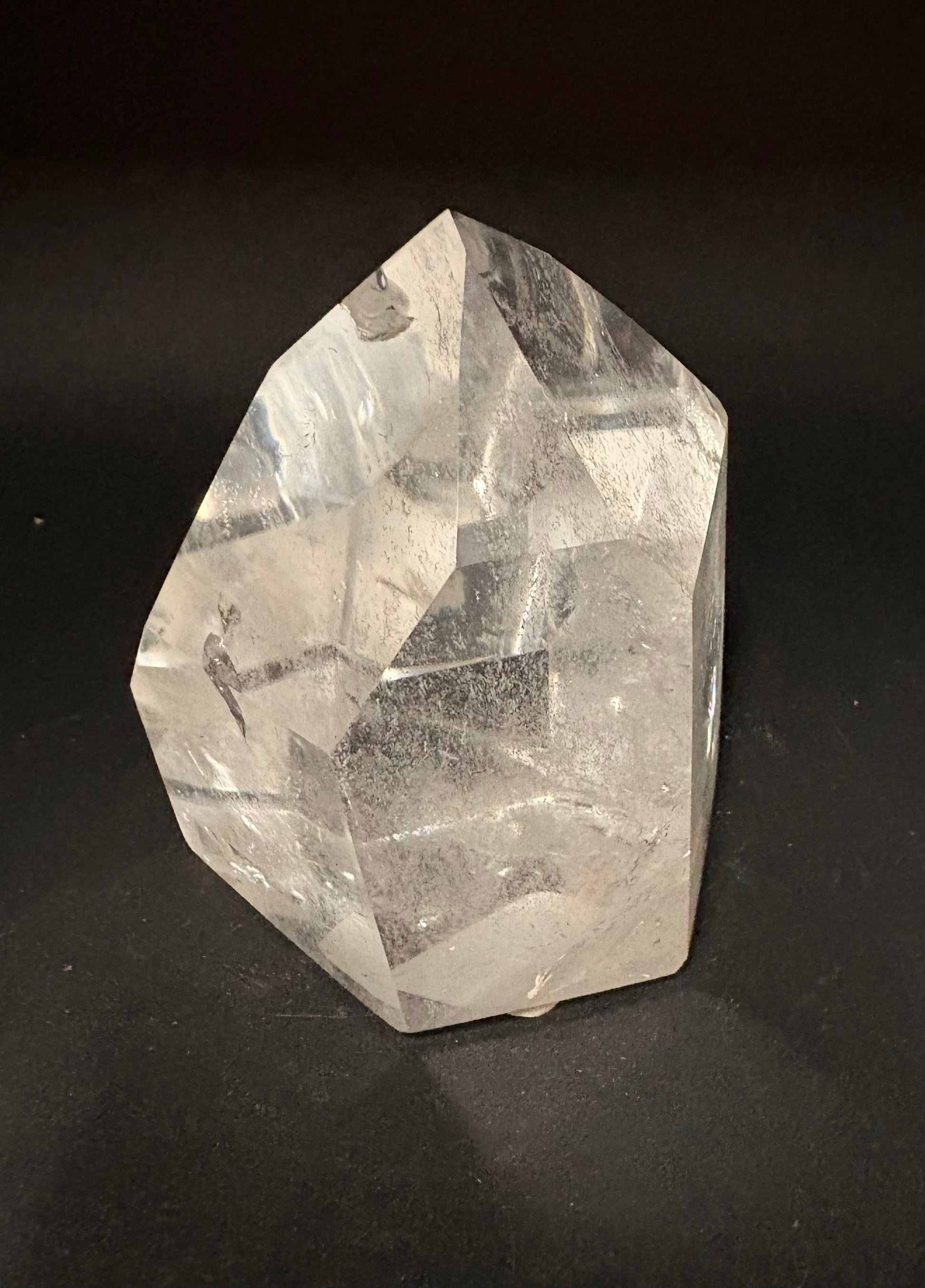 A raw, unpolished clear quartz crystal point with natural faceted edges and internal inclusions, standing upright on a dark black background.