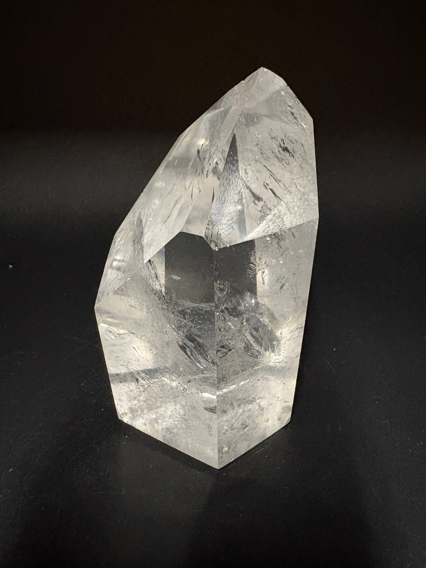A faceted clear crystal point, likely quartz, with visible internal inclusions and a pointed hexagonal shape, photographed on a dark black background.
