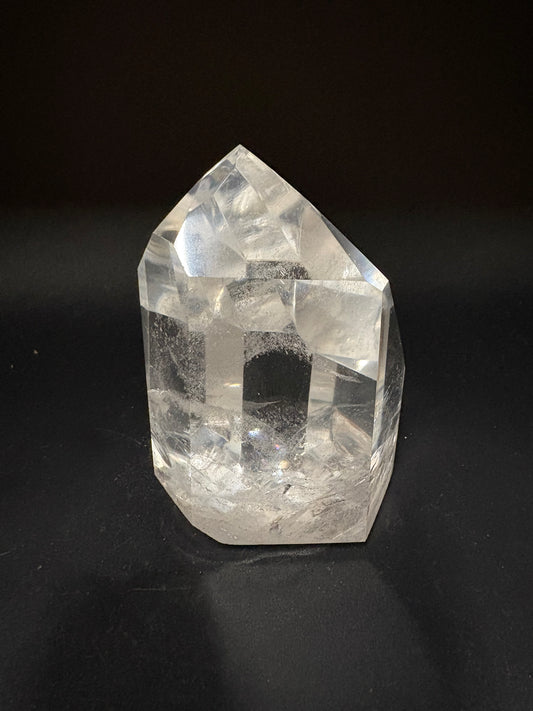 A clear quartz crystal point with natural inclusions, standing on a dark black surface against a black background.