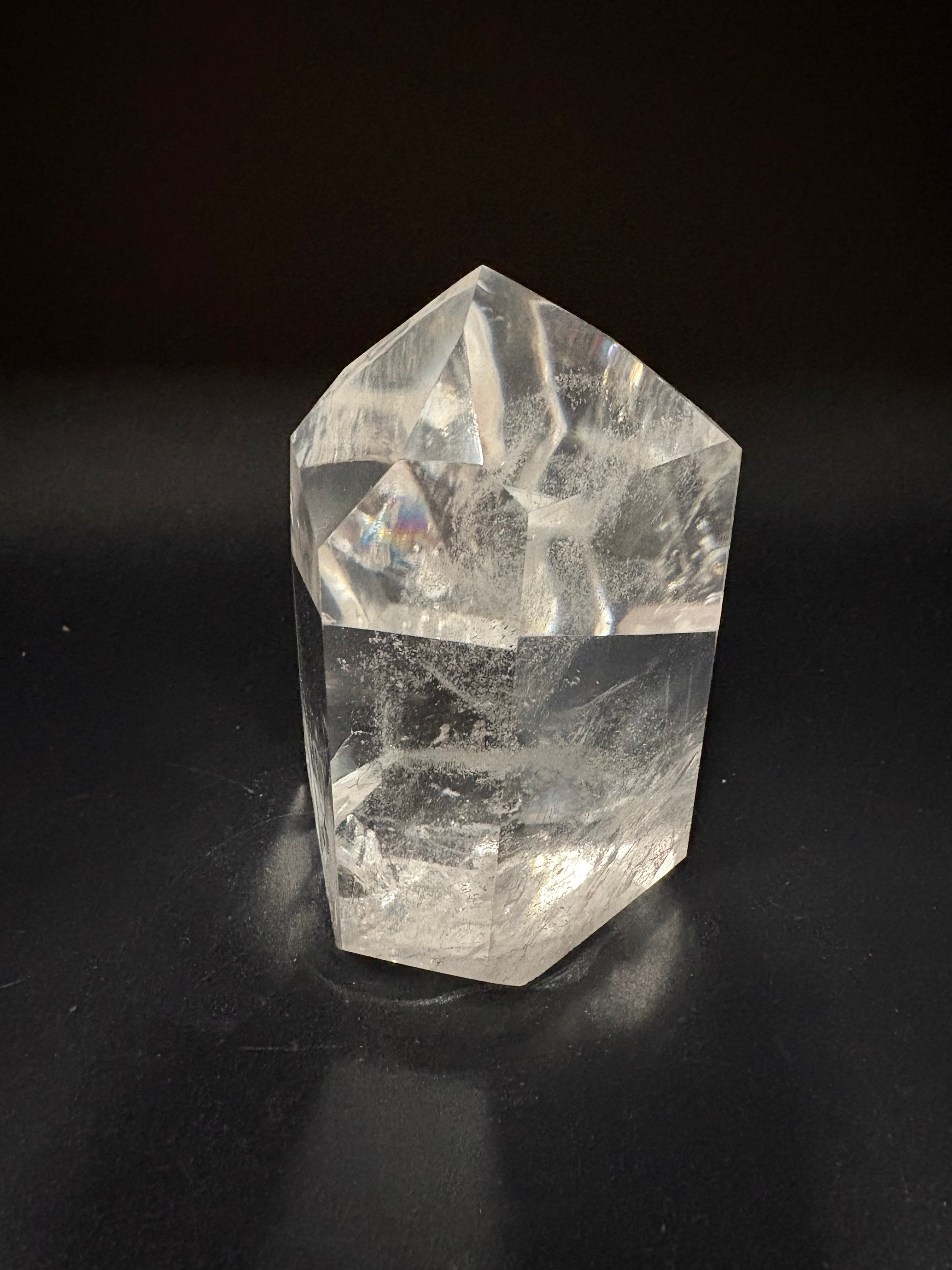 A clear quartz crystal tower with natural inclusions, standing on a dark reflective surface against a black background.