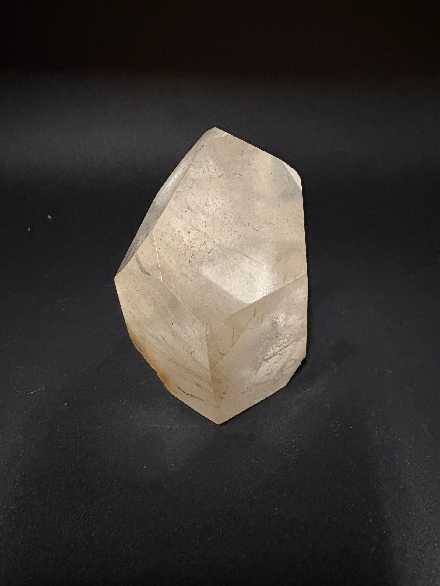 A faceted, raw crystal stone with a light beige to off-white color and subtle natural striations, resting on a dark, textured background.