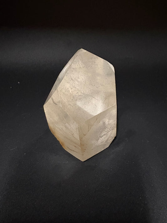 A faceted, raw crystal stone with a light beige to off-white color and subtle natural striations, resting on a dark, textured background.