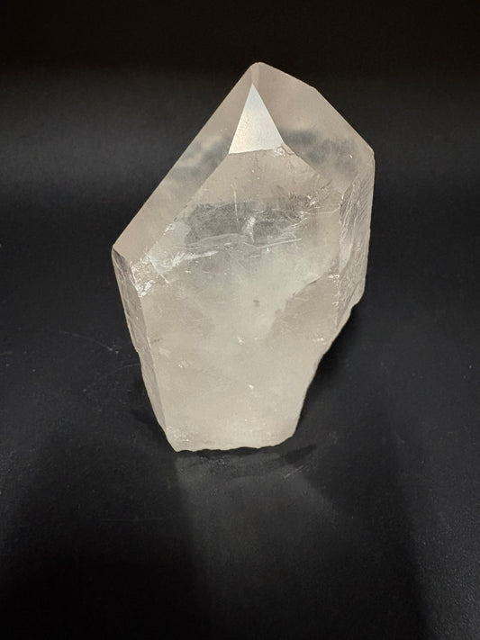 A raw, unpolished clear quartz crystal point with natural facets, displayed on a dark black background.