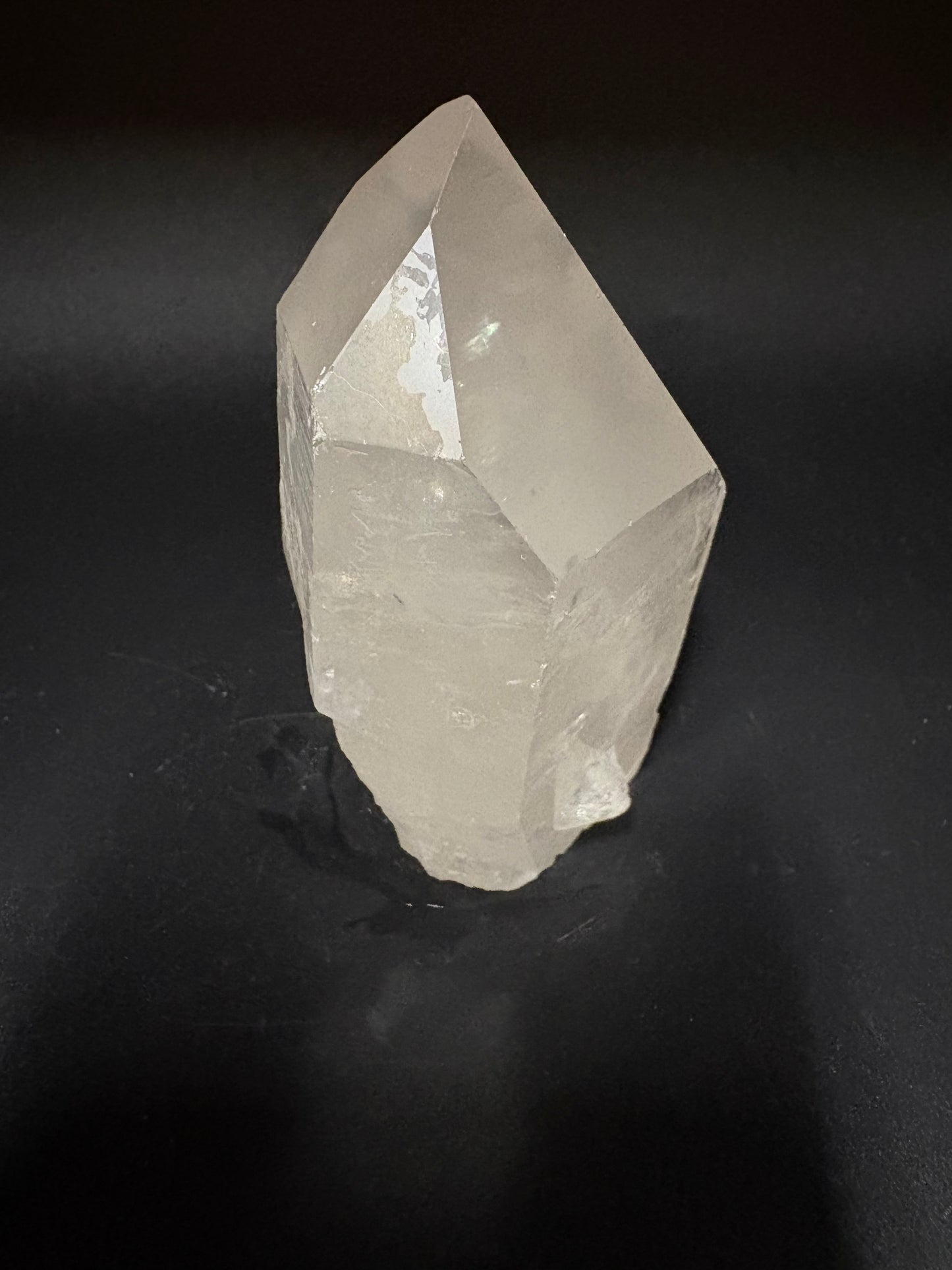 A raw, unpolished clear quartz crystal point with natural faceted edges, resting on a dark, textured black background.