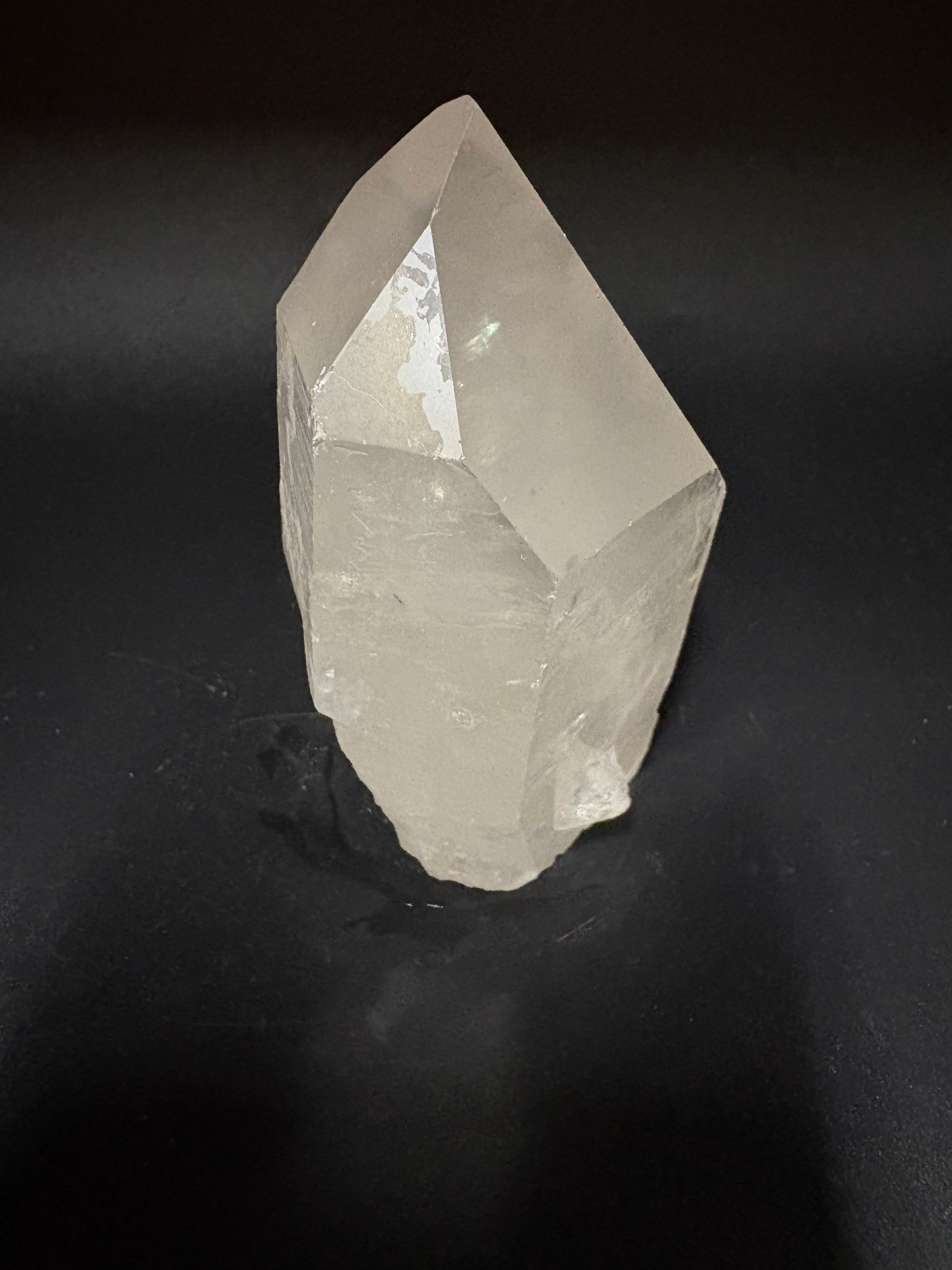 A raw, unpolished clear quartz crystal point with natural faceted edges, resting on a dark, textured black background.