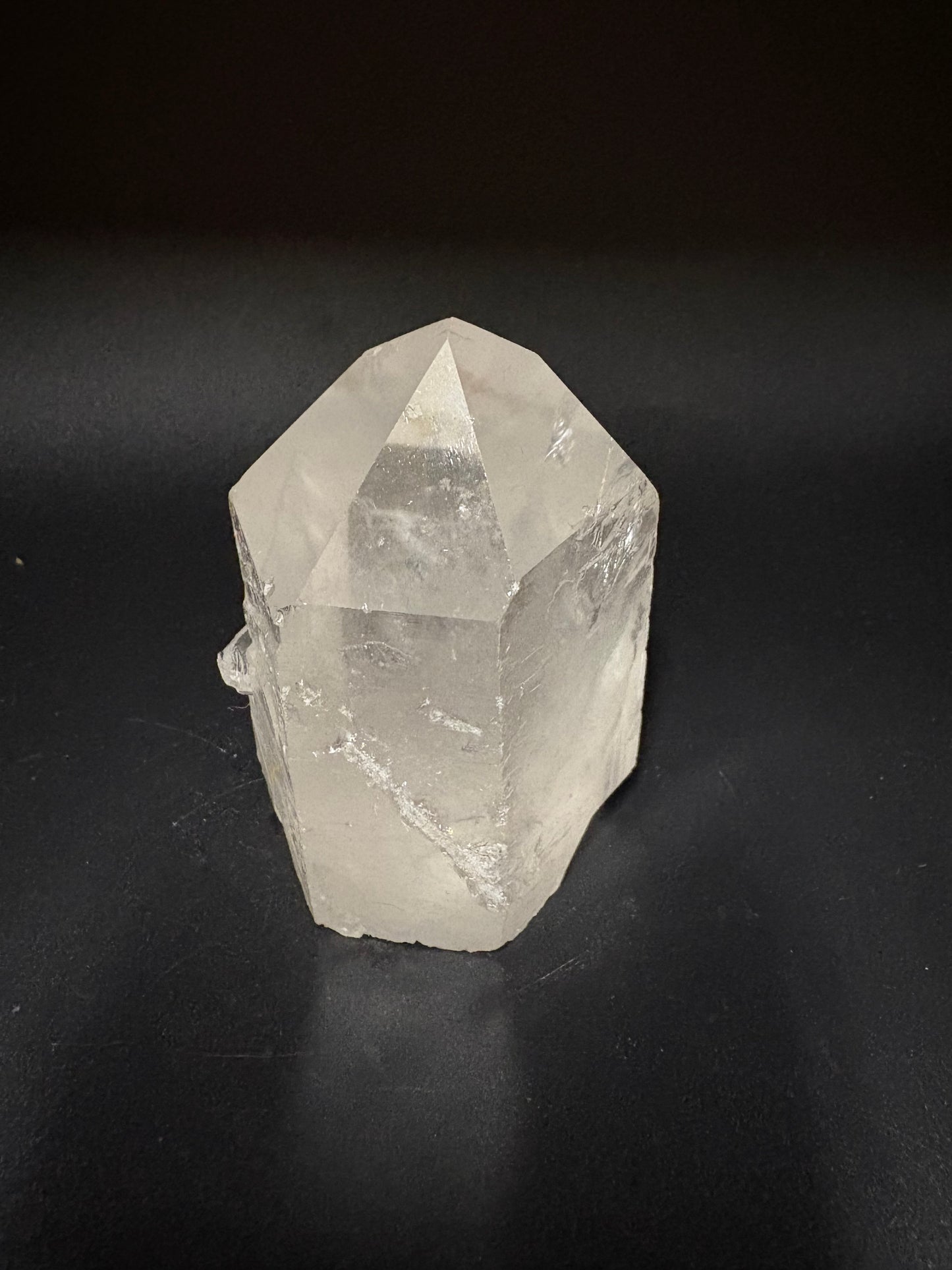 A raw, unpolished clear quartz crystal point with natural geometric facets, resting on a dark black background.