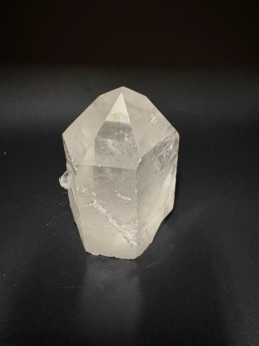 A raw, unpolished clear quartz crystal point with natural geometric facets, resting on a dark black background.