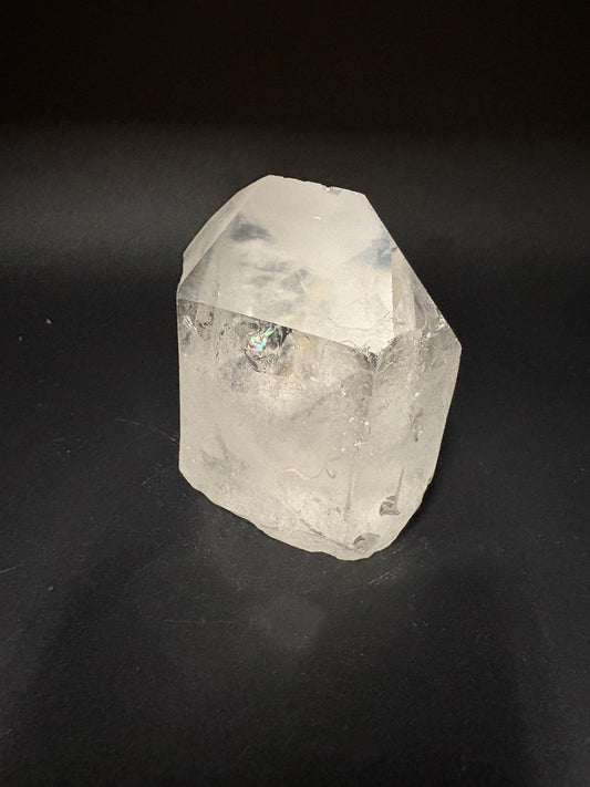 A raw, unpolished clear quartz crystal with natural facets and inclusions, resting on a dark black background.