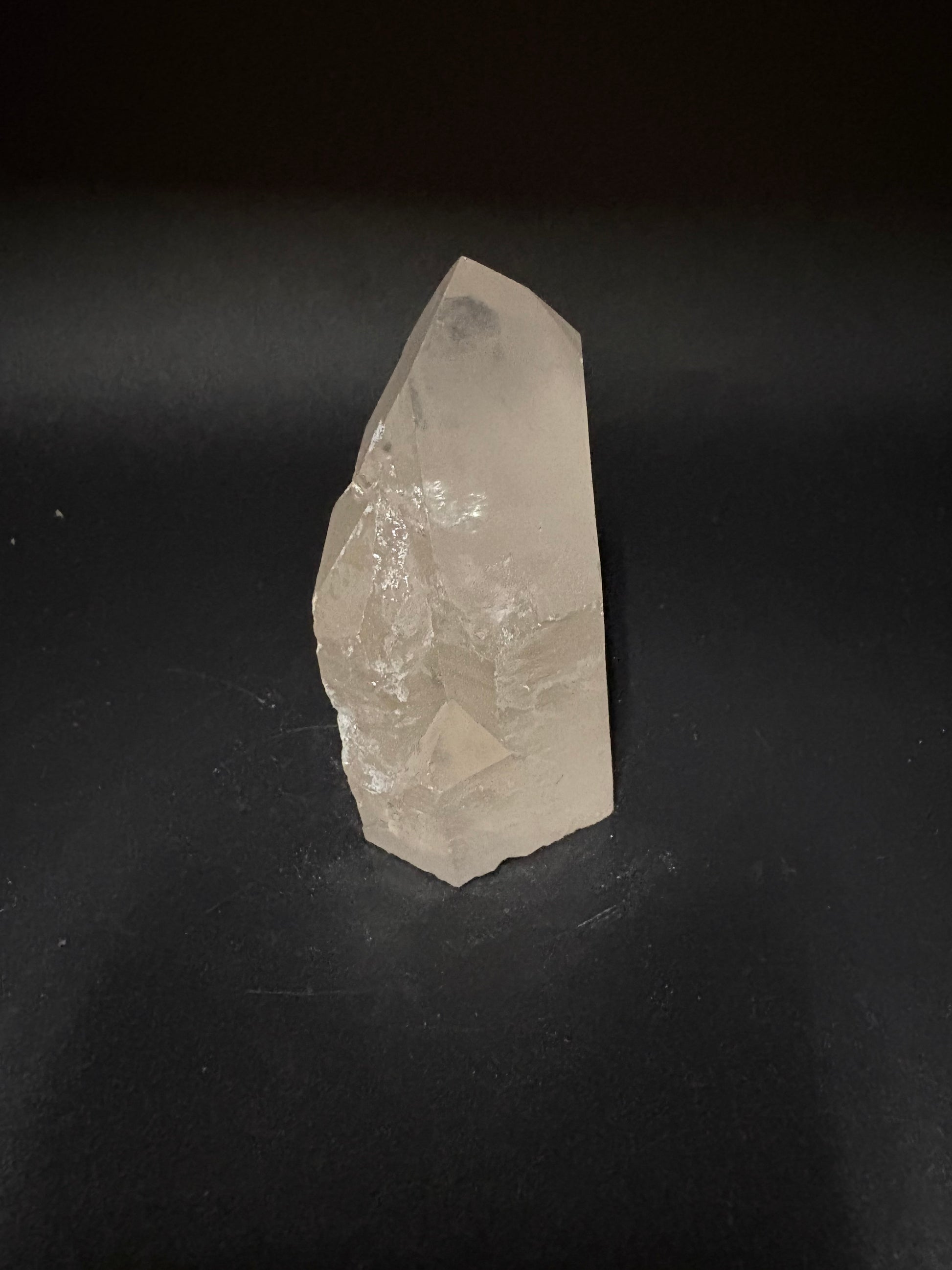 A raw, rough-cut clear quartz crystal point with natural facets, resting on a dark black background.