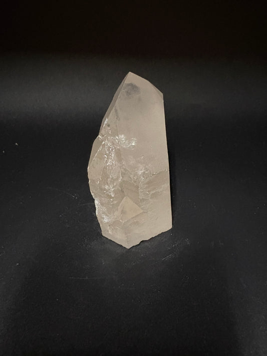 A raw, rough-cut clear quartz crystal point with natural facets, resting on a dark black background.