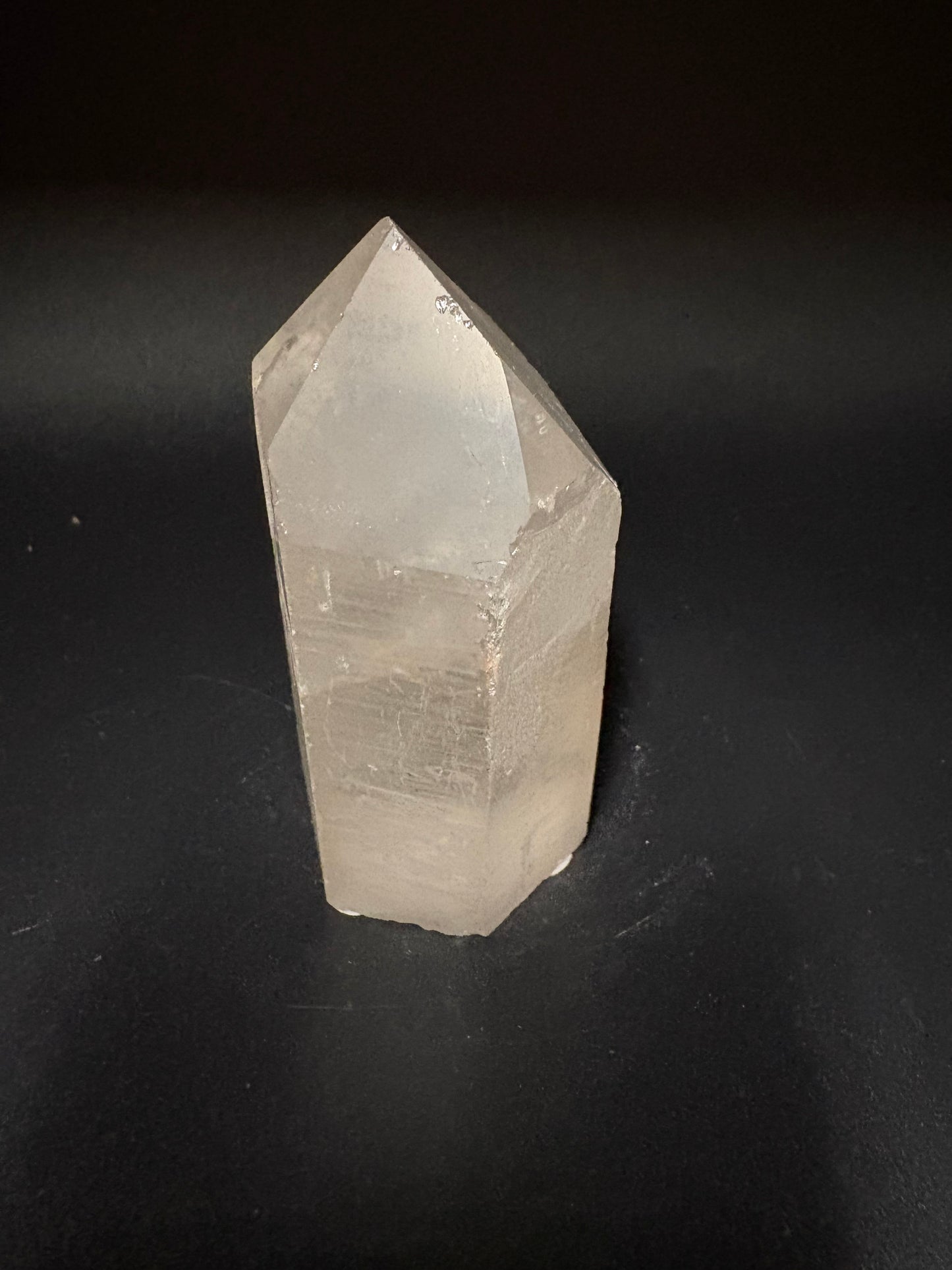A natural pointed crystal, likely quartz, with a translucent milky white appearance, resting on a dark black background.