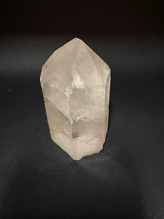 A raw, faceted clear quartz crystal point resting on a dark, matte black background.