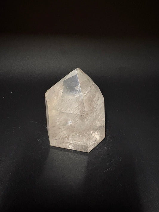 A raw, faceted clear quartz crystal point resting on a dark, matte surface.