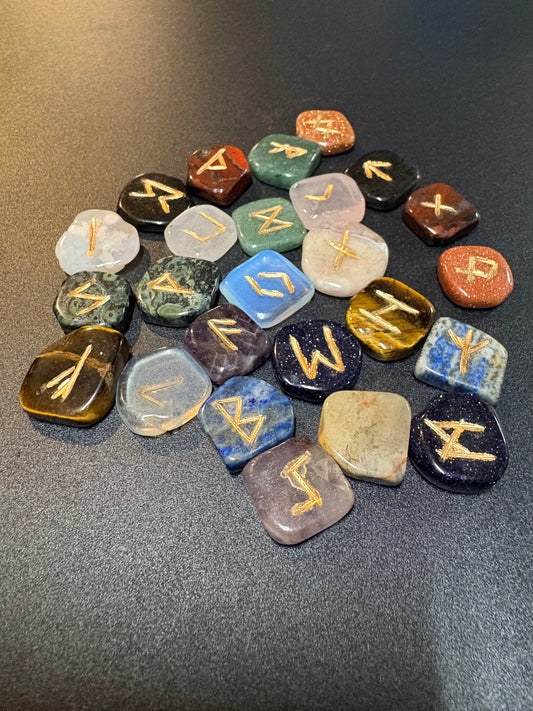 A group of polished tumbled gemstone runic tiles with engraved gold symbols, arranged in a cluster on a dark gray textured surface.