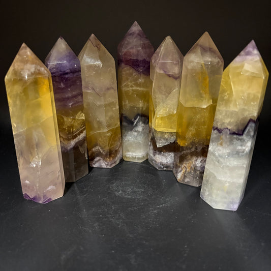 Seven natural fluorite crystal point towers of varying heights and patterns, showcasing yellow, purple, and white banding, arranged on a dark surface against a black background.