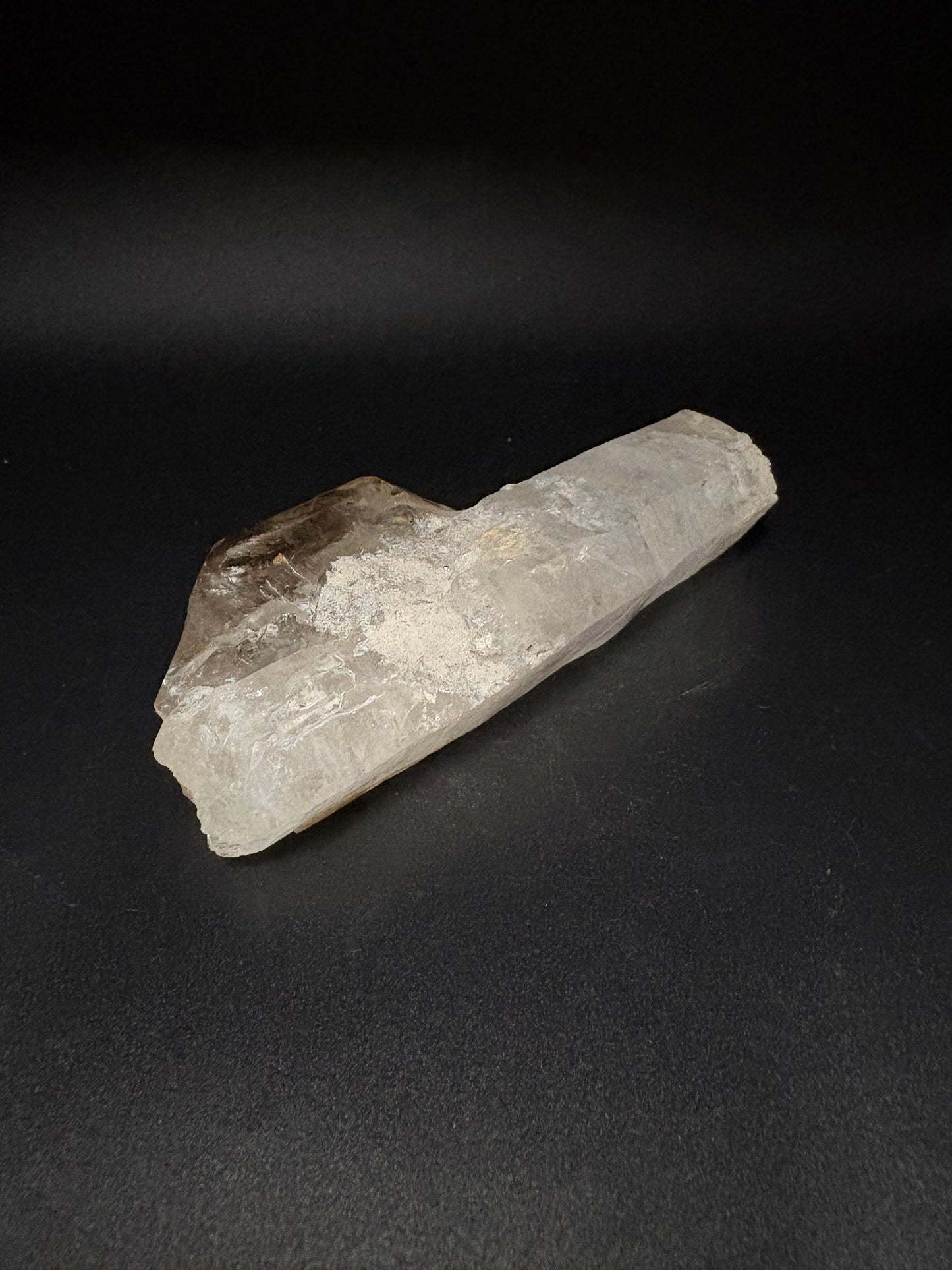 A raw, natural quartz crystal cluster with a rough, textured surface, showing a mix of translucent white quartz and dark brown inclusions, displayed diagonally on a dark black background.