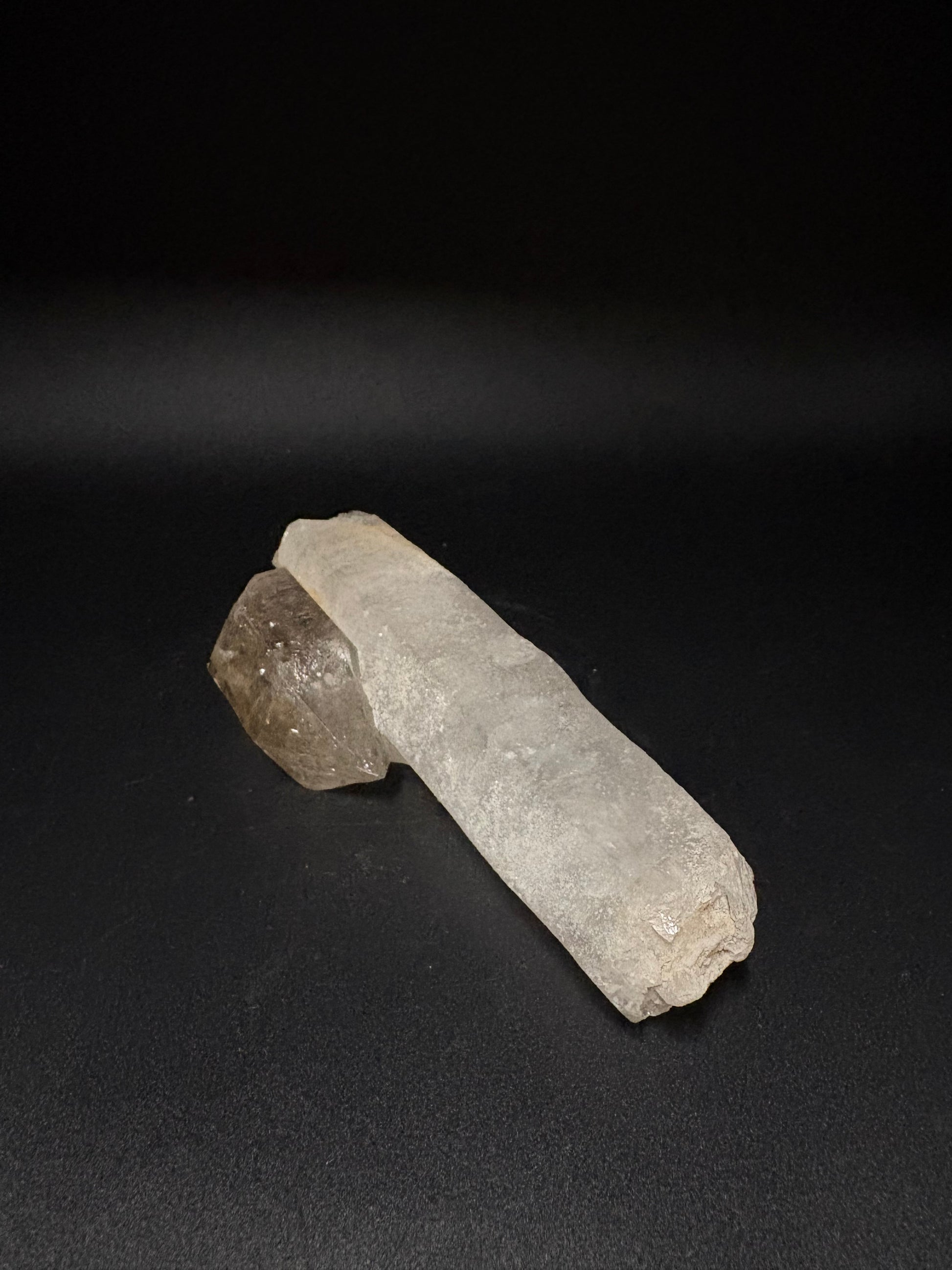 A raw crystal cluster featuring a long, pale gray translucent column and a smaller, rougher smoky brown piece, displayed on a dark black background.
