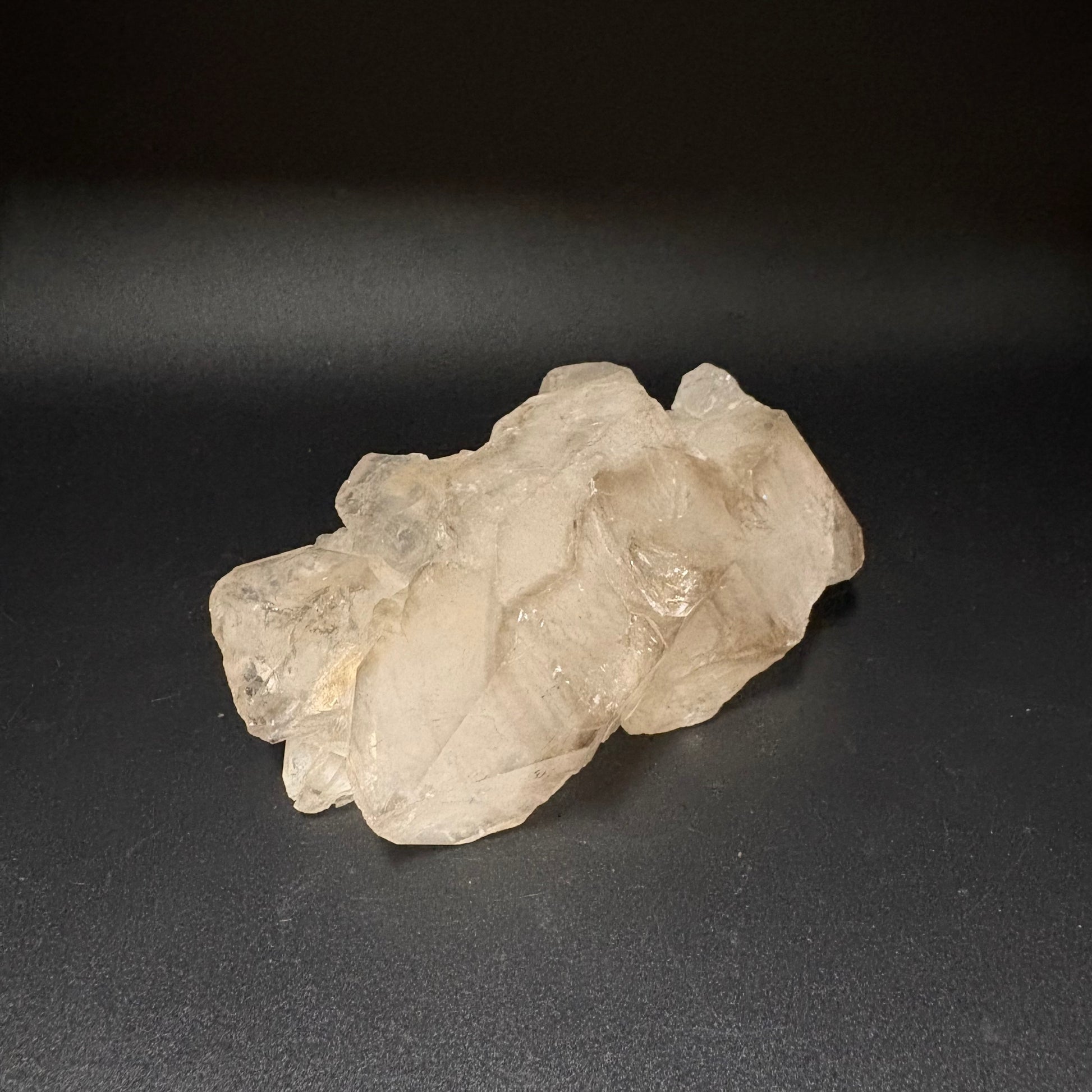 A raw, rough translucent crystal specimen, likely quartz, with natural jagged edges and internal inclusions, resting on a dark gray surface.