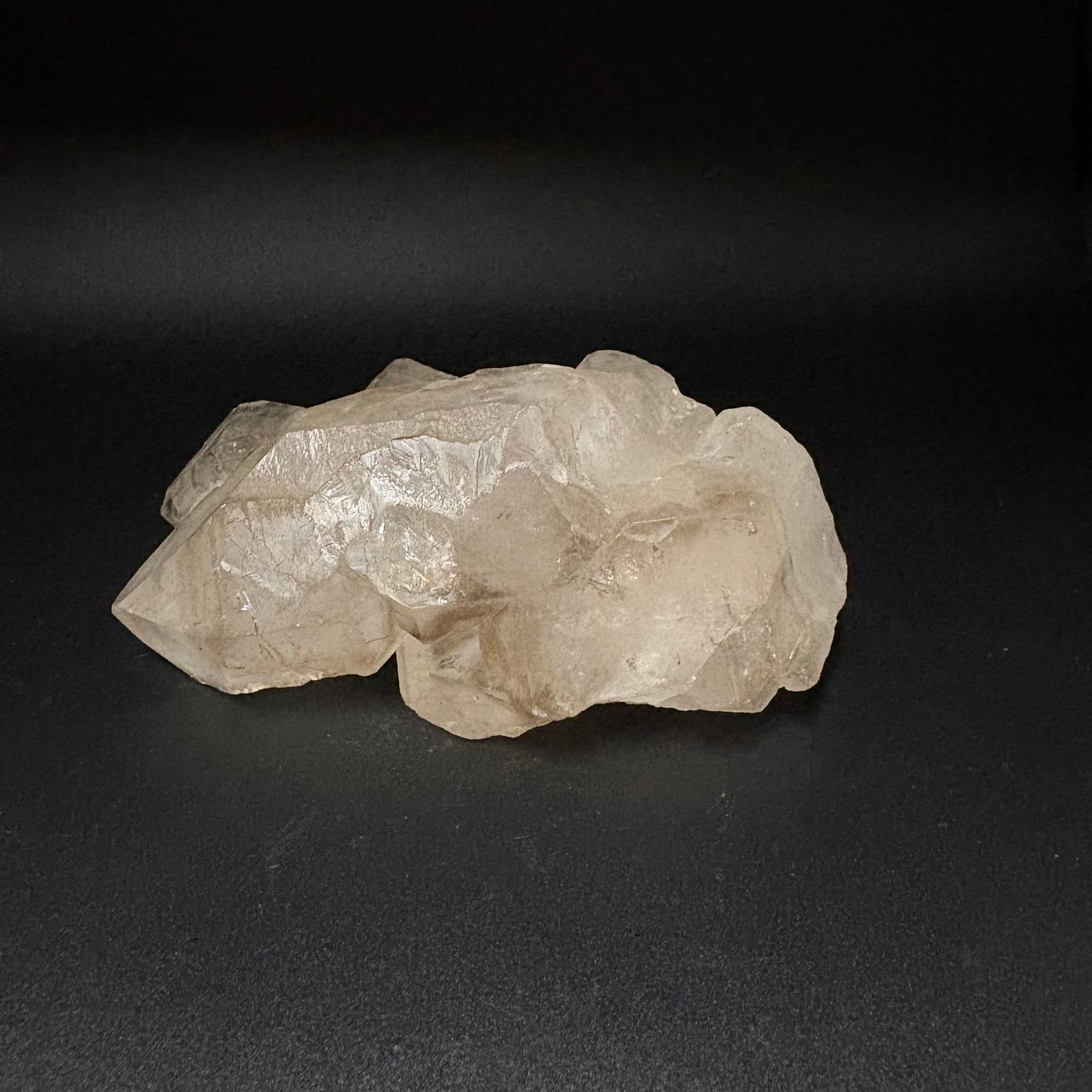 A rough, unpolished piece of clear quartz crystal with natural facets, resting on a dark gray background.
