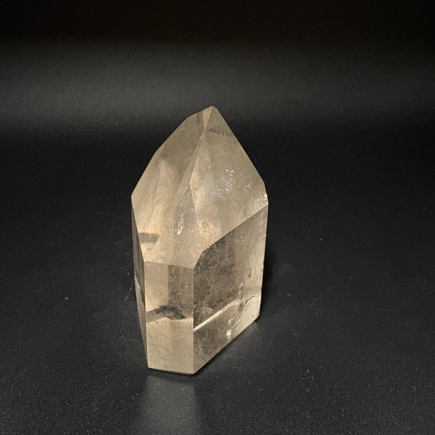 A natural clear quartz crystal point with a faceted geometric structure, standing upright on a dark, reflective surface.